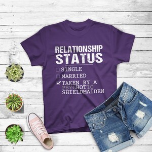 May include: Purple t-shirt with a white graphic that reads "Relationship Status" with a checkmark next to "Taken by a Psychotic Shieldmaiden" and unchecked boxes next to "Single" and "Married".