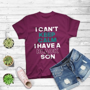 May include: A maroon t-shirt with white text that reads "I can't keep calm I have a black son" in a distressed font. The text is in teal.