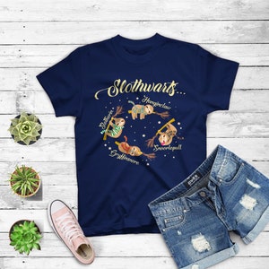 May include: A navy blue t-shirt with a gold graphic of four sloths on broomsticks. The sloths are labeled "Slytherin", "Gryffindor", "Hufflepuff", and "Ravenclaw". The text "Slothwarts..." is above the sloths.