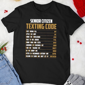 May include: Black t-shirt with white text that reads "Senior Citizen Texting Code" and a list of common senior citizen texting abbreviations and their meanings.