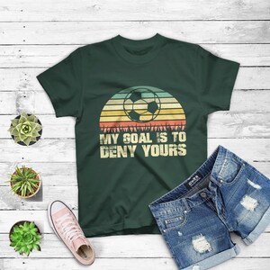 May include: A dark green t-shirt with a retro sunset graphic featuring a soccer ball. The text "My goal is to deny yours" is printed on the shirt.
