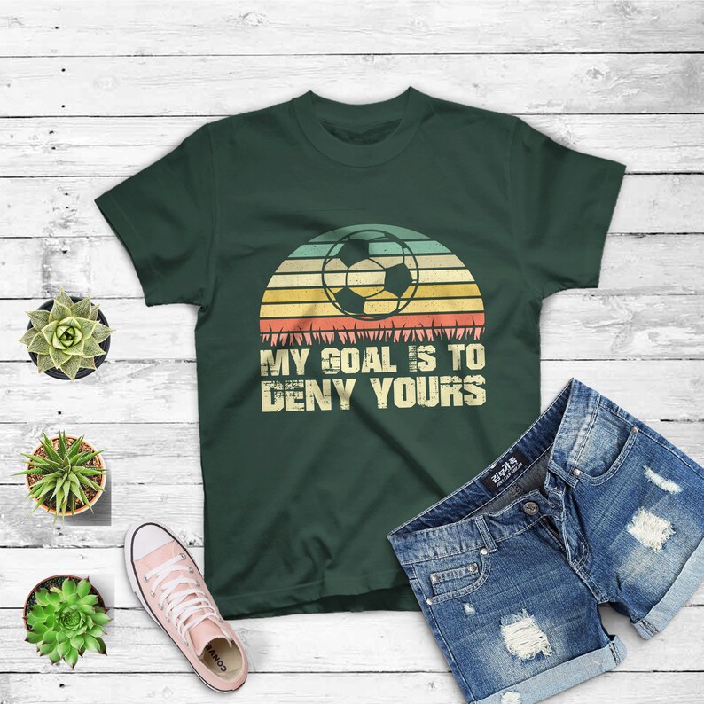 May include: A dark green t-shirt with a retro sunset graphic featuring a soccer ball. The text "My goal is to deny yours" is printed on the shirt.