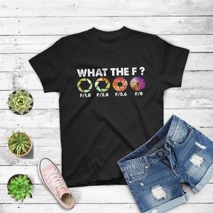 Könnte beinhalten: Schwarzes T-Shirt mit dem Text "WHAT THE F?" und vier bunten Kamera-Blenden-Diagrammen mit dem Text "F/1.8", "F/2.8", "F/5.6" und "F/8" darunter.