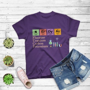 May include: Purple t-shirt with a science theme. It features the periodic table elements for Nitrogen, Erbium, Dysprosium, and Arsenic. The text "Fluorine, Uranium, Carbon, Potassium" is printed below the elements. There are also illustrations of beakers and a magnet.