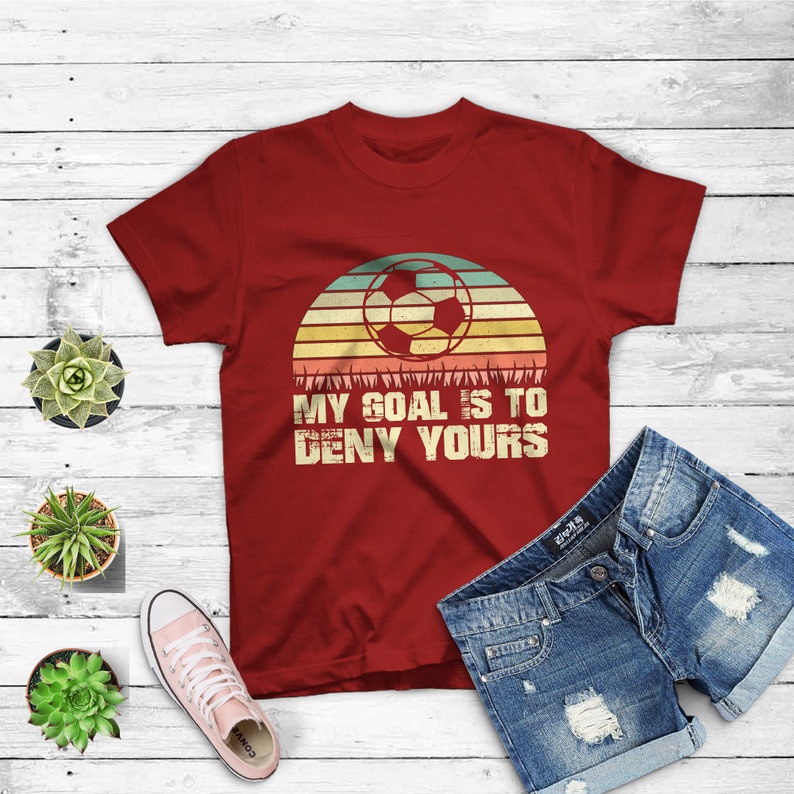 May include: Red t-shirt with a vintage-style graphic of a soccer ball in front of a sunset. The text "My goal is to deny yours" is printed below the graphic.