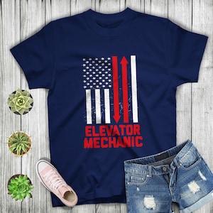May include: A navy blue t-shirt featuring a distressed American flag design and the text "ELEVATOR MECHANIC" in red.