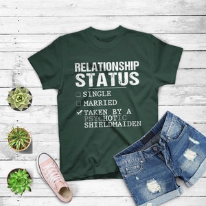May include: A dark green t-shirt with the text "RELATIONSHIP STATUS" in white. Below the text are three checkboxes with the options "SINGLE", "MARRIED", and "TAKEN BY A PSYCHOTIC SHIELDMAIDEN". The checkbox for "TAKEN BY A PSYCHOTIC SHIELDMAIDEN" is checked.