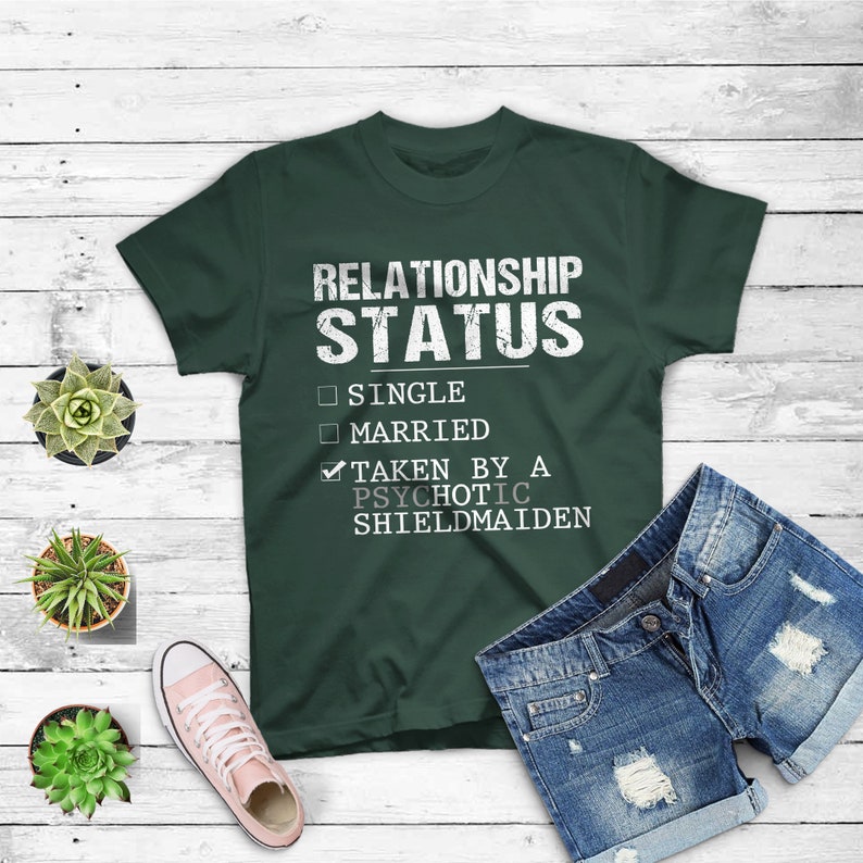 May include: A dark green t-shirt with the text "RELATIONSHIP STATUS" in white. Below the text are three checkboxes with the options "SINGLE", "MARRIED", and "TAKEN BY A PSYCHOTIC SHIELDMAIDEN". The checkbox for "TAKEN BY A PSYCHOTIC SHIELDMAIDEN" is checked.