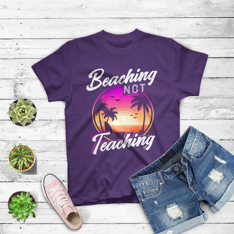 May include: A purple t-shirt with the words "Beaching Not Teaching" in white script. The design features a sunset scene with palm trees. The shirt is paired with denim shorts and pink sneakers.