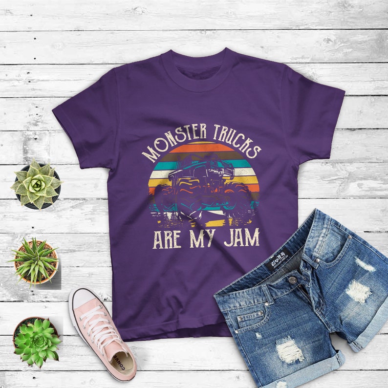 Puede incluir: Camiseta morada con un gr&aacute;fico de estilo vintage de un cami&oacute;n monstruo conduciendo a trav&eacute;s de una puesta de sol. El texto "Monster Trucks Are My Jam" est&aacute; impreso en la camiseta.
