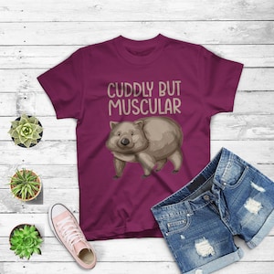 Könnte beinhalten: Ein maroonfarbenes T-Shirt mit einem Cartoon-Wombat und dem Text "CUDDLY BUT MUSCULAR".