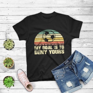 May include: Black t-shirt with a vintage-style graphic of a soccer ball in front of a sunset. The text "My goal is to deny yours" is printed below the graphic.