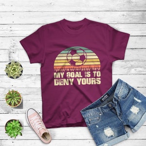 May include: A maroon t-shirt with a vintage-style graphic of a soccer ball in front of a sunset. The text "My goal is to deny yours" is printed below the graphic.