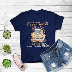 May include: Navy blue t-shirt with a graphic of a stack of books, a cup of tea, and fall leaves. The text "That's what I do I read books I drink tea and I know things" is printed on the shirt.