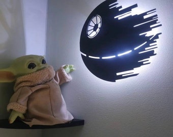 Könnte beinhalten: Eine schwarze Death-Star-Wandleuchte mit weißer LED-Beleuchtung. Ein grünes Baby-Yoda-Plüschtier befindet sich auf einem schwarzen Regal und greift nach dem Licht. Die Wand ist hellgrau gestrichen.