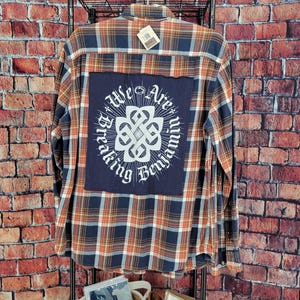 May include: A long-sleeved flannel shirt with a plaid pattern in shades of blue, orange, and brown. The back features a navy blue patch with the words "We Are Breaking Benjamin" in white, surrounding a white emblem. The shirt is hanging on a black metal rack.