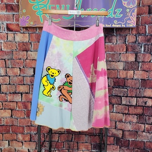 May include: A colorful patchwork skirt with a pink waistband and a-line silhouette. The skirt features panels of blue, tie-dye, and pink fabric, with two cartoon bear graphics. The skirt is hanging on a wooden hanger.