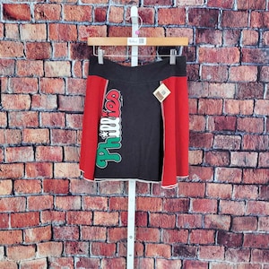 May include: A black and red skirt with the word "Phillies" in green and white script. The skirt has a white trim and is hanging on a hanger against a brick wall.