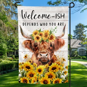 May include: A garden flag with a white wooden background featuring a brown Highland cow with a sunflower crown. The text on the flag reads "Welcome-ish Depends Who You Are".