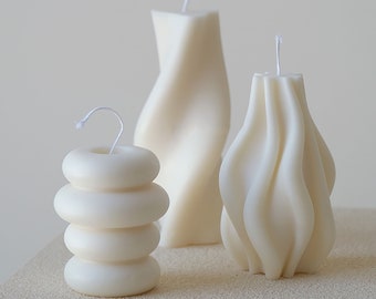 May include: Three white candles with unique shapes and textures. The candles are arranged on a white surface. The candle on the left is a stack of four rings, the candle in the center is a tall, wavy candle, and the candle on the right is a shorter, wavy candle.
