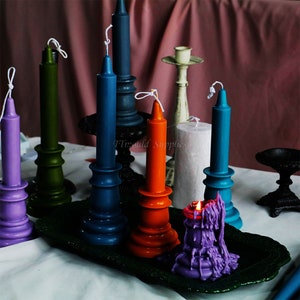 May include: A collection of colorful candles on decorative candle holders. The candles are in shades of purple, green, blue, orange, and teal. The candle holders are made of metal and have a vintage look. The candles are arranged on a dark green tray.