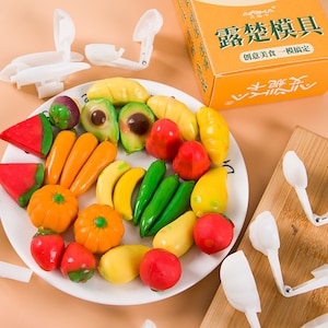 May include: A white plate with a variety of colorful fruit-shaped candies. The candies are made to look like watermelon, avocado, banana, carrots, peppers, and more. The plate is surrounded by white plastic molds for making the candies.