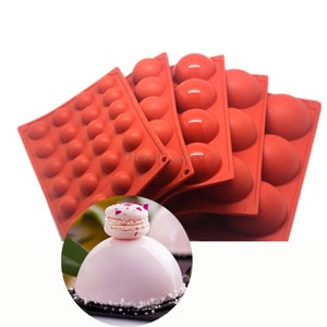 Puede incluir: Conjunto de cinco moldes de silicona rojos para hacer pasteles o postres redondos. Los moldes están apilados uno encima del otro. El molde más grande tiene 24 cavidades redondas individuales. Los moldes más pequeños tienen 6 cavidades redondas individuales. Un pastel rosa con un macaron en la parte superior se muestra en primer plano.