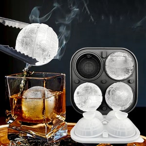 May include: A black and white ice cube tray with four round molds shaped like the Death Star from Star Wars. The tray is made of silicone and has a white base. There are four ice cubes in the tray, and one is being lifted out with tongs.