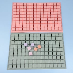 May include: Two silicone moulds for making small square-shaped sweets or chocolates. The top mould is pink and the bottom mould is green. There are several small, colourful sweets in the green mould.