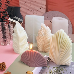 May include: Four different candle moulds, two leaf-shaped, one fan-shaped, and one cylindrical. The leaf-shaped moulds are made of white silicone and the fan-shaped mould is purple. The cylindrical mould is white and has a smooth surface. The moulds are on a pink and white surface.