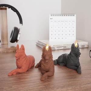 Puede incluir: Tres velas con forma de perro pastor alemán, una marrón, una negra y una marrón claro, con llamas encendidas. Las velas están sobre una superficie de madera.
