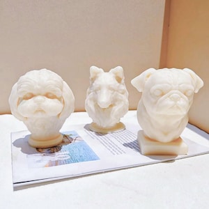 Könnte beinhalten: Drei cremefarbene Hundeskulpturen. Die Skulpturen zeigen einen Shih Tzu, einen Collie und eine Dogge. Der Shih Tzu und die Dogge sind Büsten, während der Collie ein Kopf ist. Die Skulpturen stehen auf einer Zeitung.