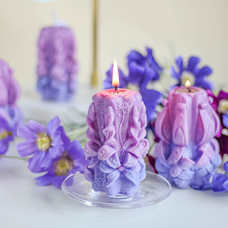 May include: Three purple and pink decorative candles with intricate floral designs. The candles are on a white surface with purple flowers.