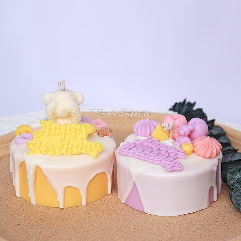May include: Two decorated birthday cake candles. The candle on the left is yellow with white icing and a white teddy bear on top. The candle on the right is purple with white icing and a variety of colorful decorations. Both candles have the words "Happy Birthday" written on them.