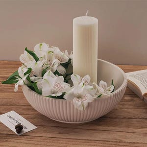 May include: White ceramic bowl with a ribbed design, filled with white flowers and a white pillar candle. The bowl is sitting on a wooden surface.