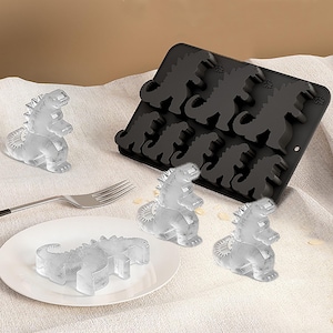 May include: A black silicone dinosaur-shaped ice cube tray with several dinosaur ice cubes. The ice cubes are on a white plate and on a light-colored surface. A fork is also present.