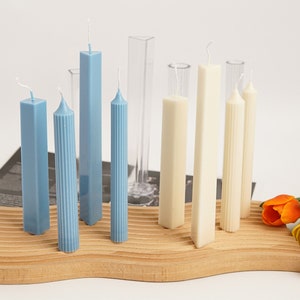 Puede incluir: Surtido de velas pilares de diferentes formas y colores, incluyendo azul y blanco, expuestas sobre una superficie de madera. Las velas están hechas con una textura acanalada y un acabado liso.