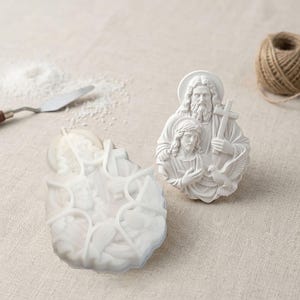 May include: A white silicone mold featuring a detailed religious relief of two figures, one holding a cross and the other with a dove, alongside a finished white plaster casting of the same design, showcasing intricate facial features and robes.