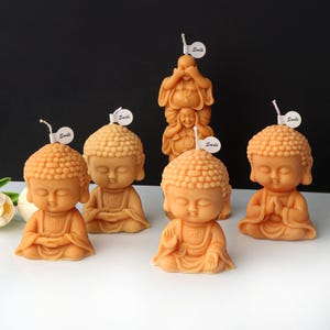 May include: Five small, hand-crafted, beige Buddha candles with a wick on top. Each candle has the word "Smile" written on a small white label.