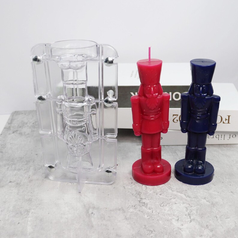 May include: A clear plastic mold and two nutcracker-shaped candles. One candle is red, the other is dark blue. The candles are standing upright. The mold is transparent and has a nutcracker shape.