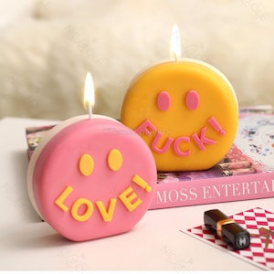 Può includere: Due candele a forma di faccina sorridente, una gialla con la parola "FUCK!" e una rosa con la parola "LOVE!" su un libro rosa e bianco.