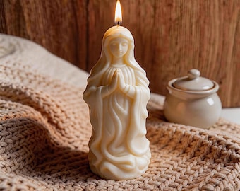 May include: A cream-colored candle shaped like a praying figure with a lit wick. The candle is placed on a beige knitted surface, with a small ceramic jar in the background. The candle's flame is bright against the soft background.