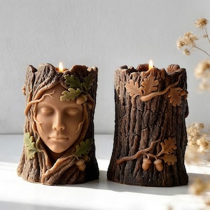 May include: Two brown tree trunk candles with a face carved into one and oak leaves and acorns on the other. The face candle has a woman's face with closed eyes and green leaves. Both candles have lit flames.