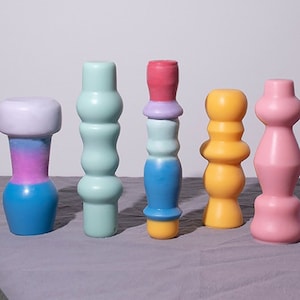 以下が含まれることがあります： 異なる形とデザインのカラフルなセラミックの花瓶が5つあります。花瓶は灰色の表面に一列に並んでいます。花瓶は滑らかで光沢のあるセラミックでできており、ピンク、ブルー、グリーン、イエロー、パープルなど、さまざまな色があります。
