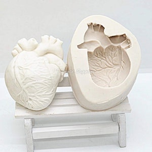 May include: A white silicone mould of a human heart. The mould is open and shows the detailed anatomical features of the heart. The mould is sitting on a white wooden bench.