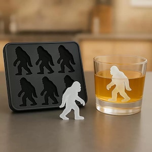 May include: Black silicone ice cube tray with six Bigfoot-shaped molds. A clear glass of amber liquid contains a Bigfoot-shaped ice cube. A single Bigfoot-shaped ice cube sits on the surface. The ice cube tray and ice cubes are on a gray surface.