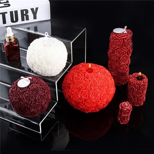 May include: A collection of rose-shaped candles in various sizes and colors. The candles are white, burgundy, and red, with a detailed rose pattern. Some candles have a lit wick, and all have a small tag attached.