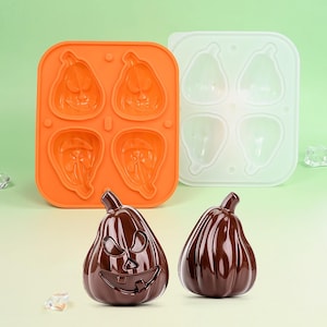 Könnte beinhalten: Eine orangefarbene Silikonform mit vier kürbisförmigen Hohlräumen, neben einer weißen Form mit vier Kürbisformen. Zwei dunkelbraune, kürbisförmige Süßigkeiten liegen vor den Formen, eine mit einem geschnitzten Gesicht.