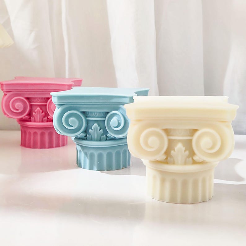 K&ouml;nnte beinhalten: Drei dekorative ionische S&auml;ulenskulpturen in Rosa, Blau und Creme. Die S&auml;ulen weisen detaillierte Schn&ouml;rkel und kannelierte Sch&auml;fte auf, die an klassische Architektur erinnern. Die Skulpturen sind auf einer wei&szlig;en Oberfl&auml;che angeordnet.