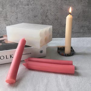 Puede incluir: Un molde de silicona transparente para hacer velas, con seis cavidades redondas. Se muestran tres velas rosas y una vela blanca junto al molde. La vela blanca está encendida.